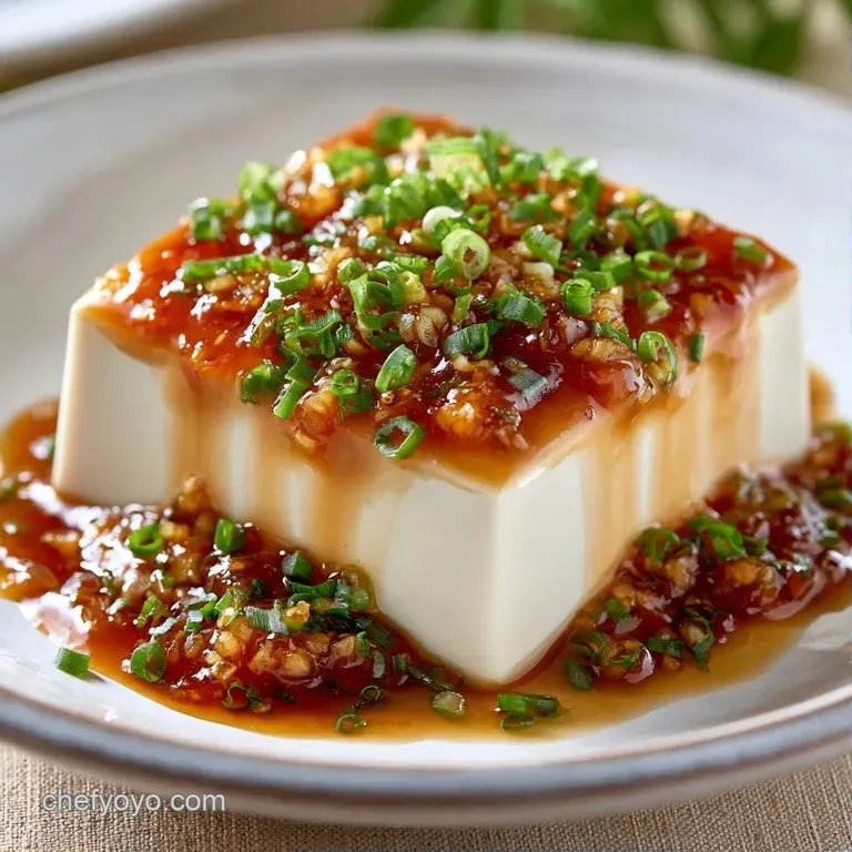 A smooth block of white tofu on a minimalist plate, drizzled with dark sauce and sprinkled with fresh green herbs.